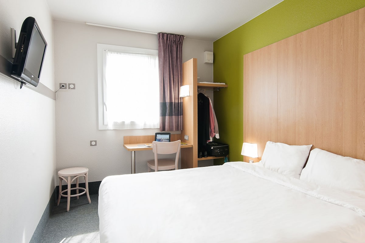 Une chambre d’hôtel avec un lit, un bureau avec un ordinateur portable, une chaise, un téléviseur fixé au mur et une fenêtre avec des rideaux.