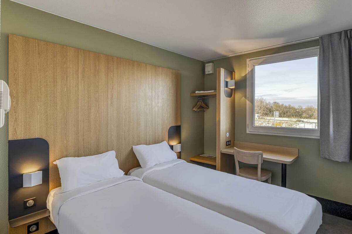 Une chambre d’hôtel avec deux lits, un bureau, une chaise, une fenêtre donnant sur un paysage agréable et une tête de lit en bois.