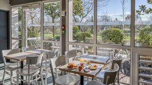 A dining area with tables and chairs, large windows with a view of trees and a fence, and a fire extinguisher on the wall.