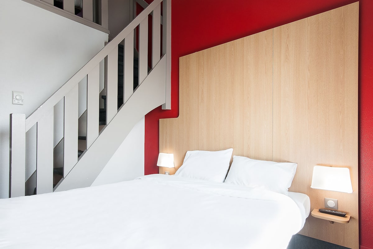 Une chambre d’hôtel avec un lit, deux lampes de chevet, une tête de lit en bois et un escalier blanc.