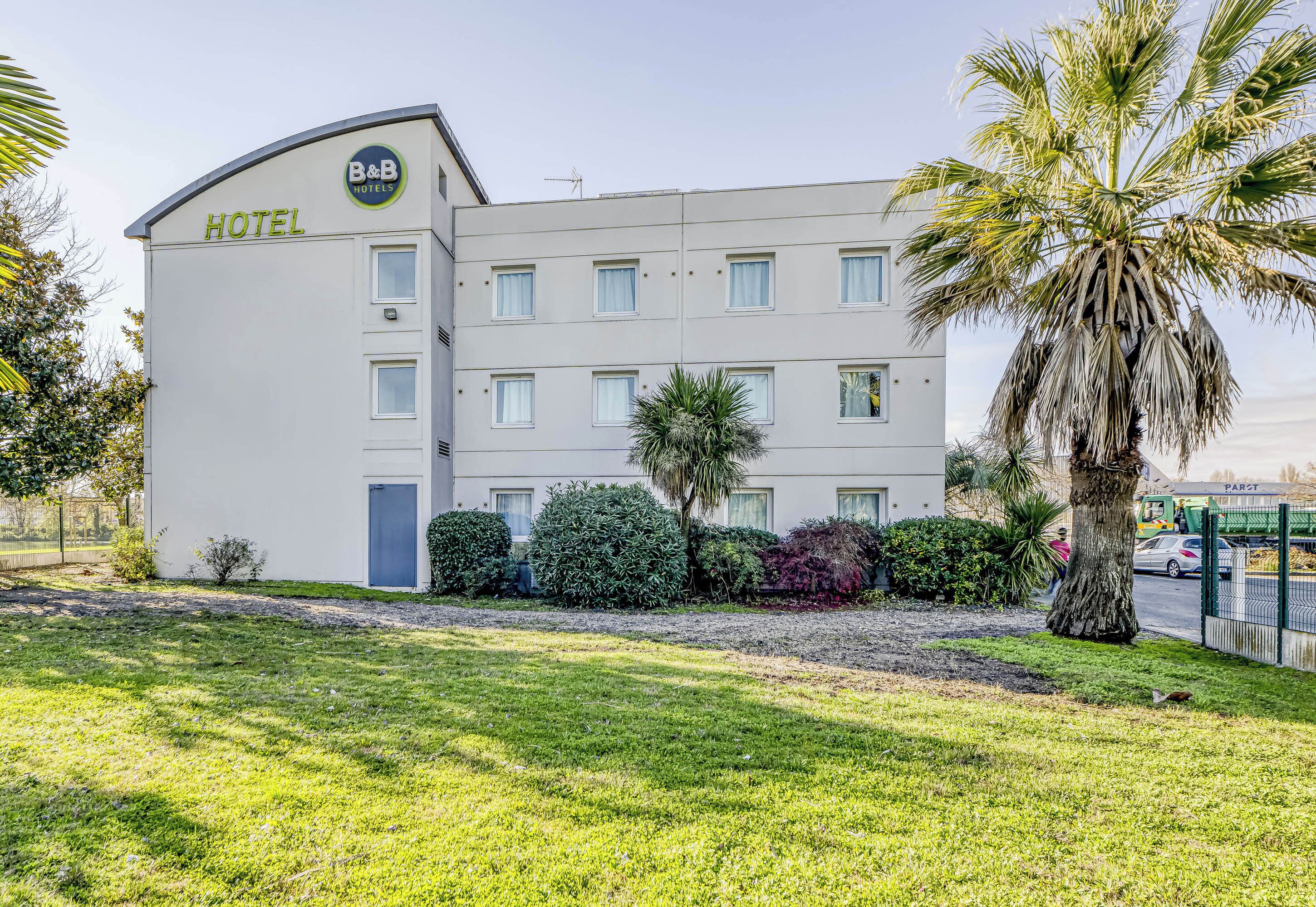 A B&B hotel with a modern design, featuring a curved facade, multiple windows, and a well-maintained lawn with palm trees.