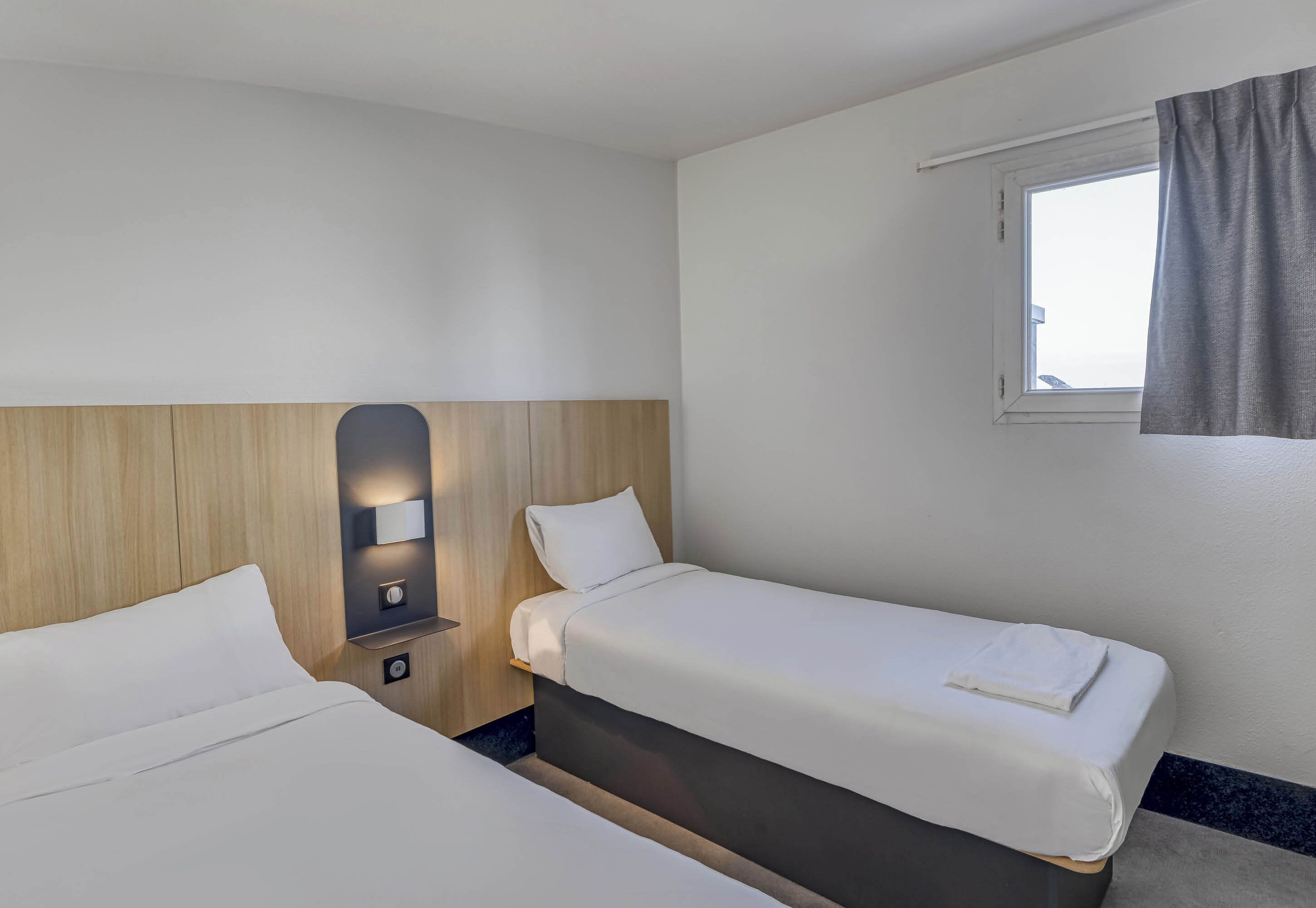 A hotel room with two beds, a bedside table with a lamp, a window with curtains, and a view of the sky.