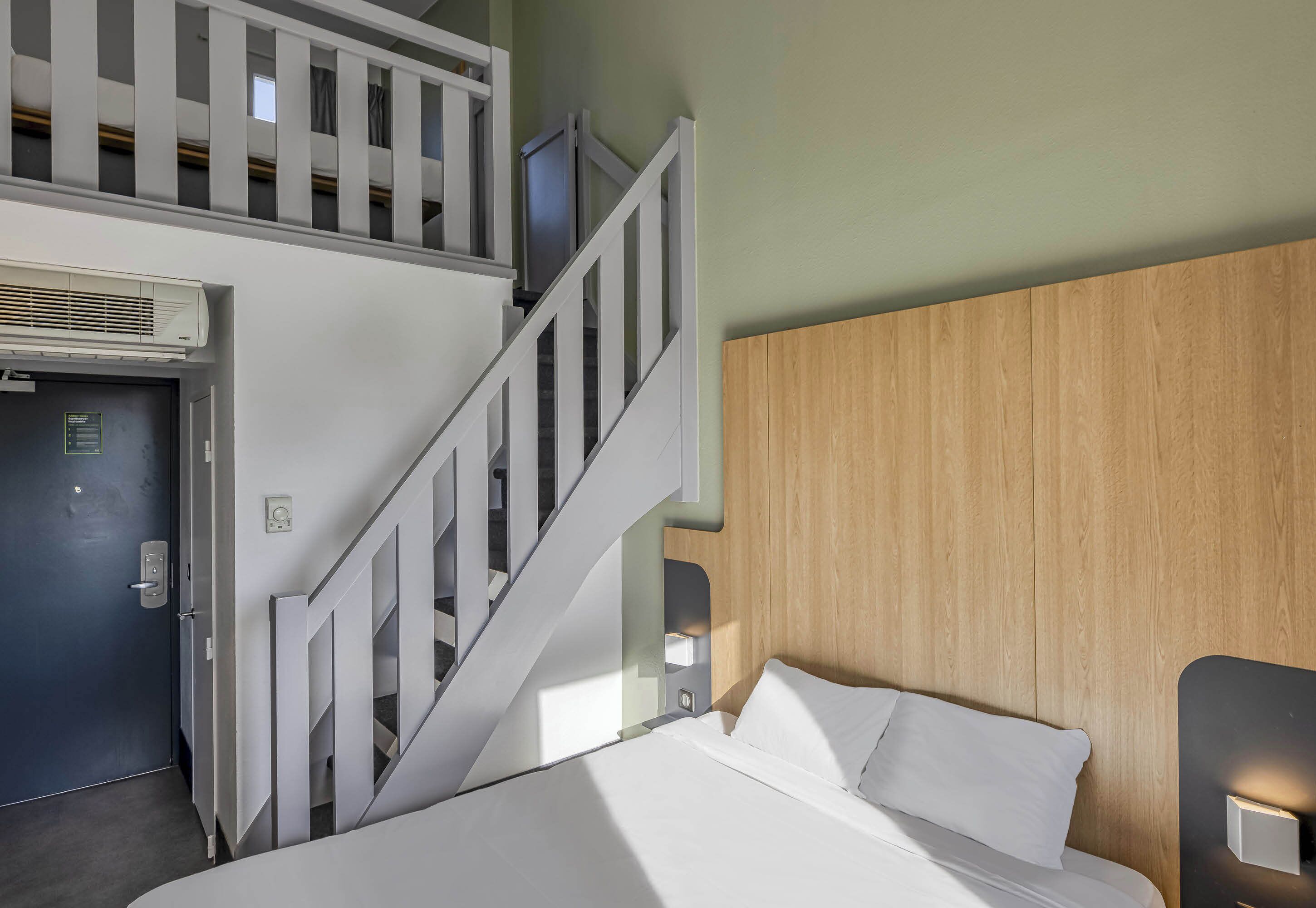 Habitación de hotel con cama, cabecera de madera, escalera y puerta con unidad de aire acondicionado.
