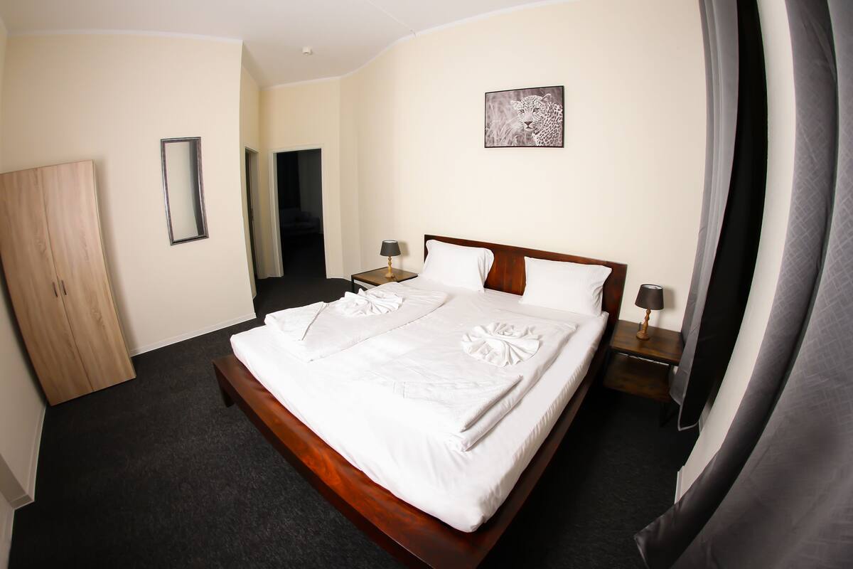 A neatly made bed with a wooden headboard, a bedside table with a lamp, a mirror on the wall, and a leopard artwork.
