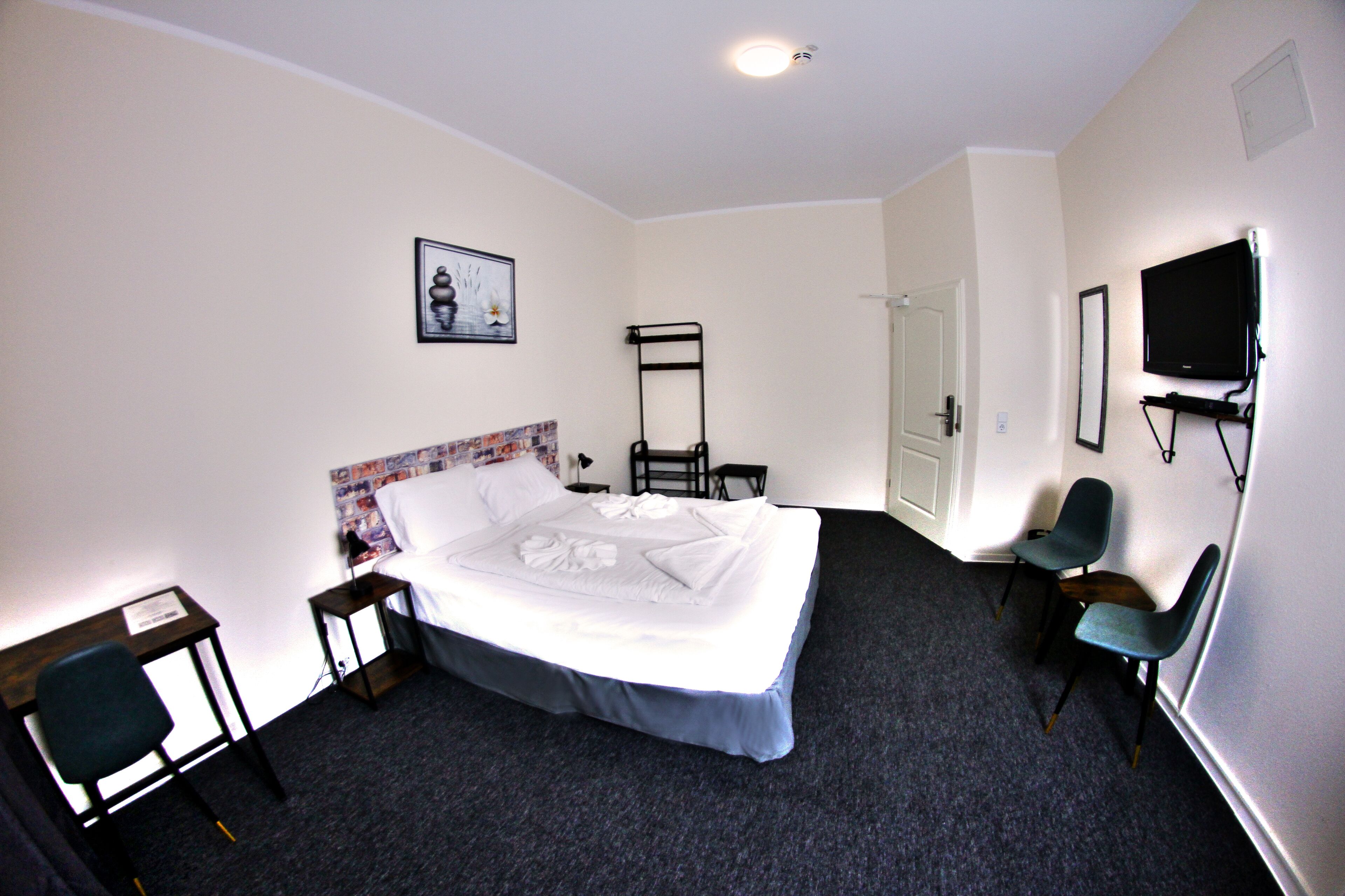 A hotel room with a neatly made bed, a desk with a chair, a TV mounted on the wall, and a door leading to another room.