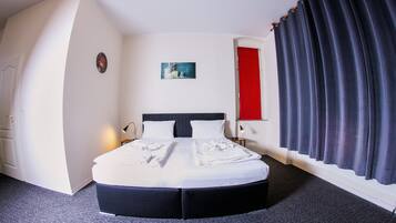 A neatly made bed in a hotel room with a dark-colored headboard, a red door, and a dark curtain.