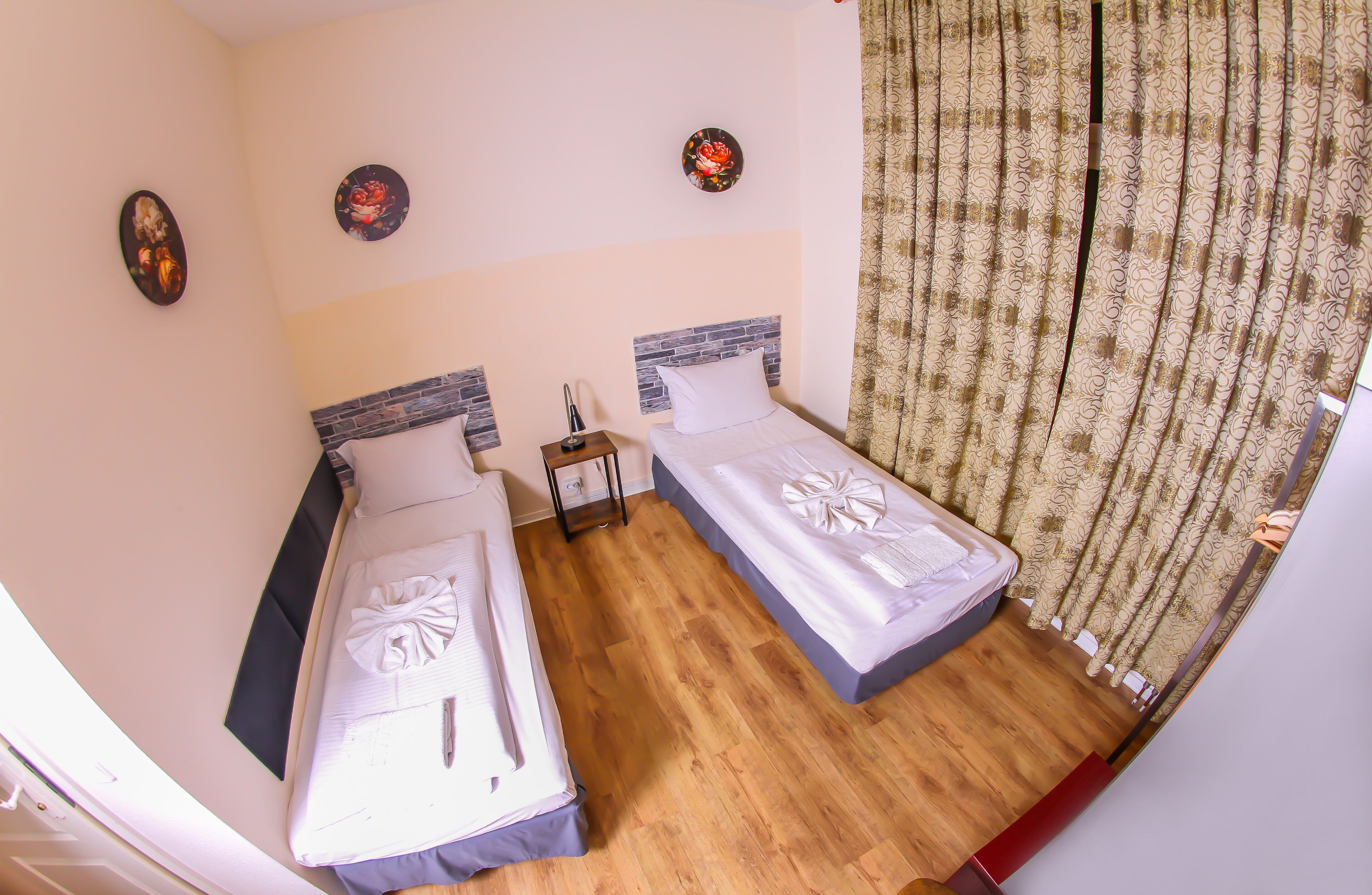 A hotel room with two beds, wooden flooring, a small bedside table with a lamp, and a curtain.