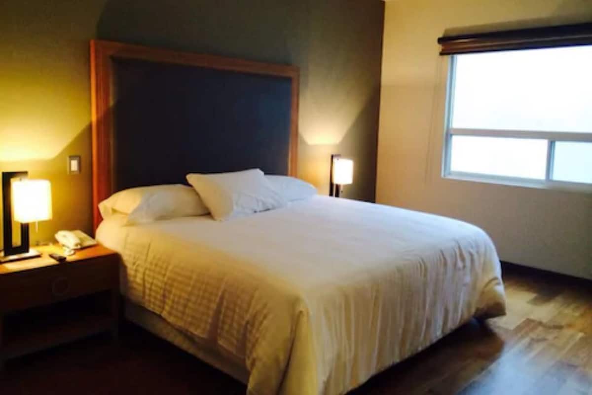 Una habitación de hotel con una cama grande, dos mesitas de noche con lámparas y una ventana con persianas.