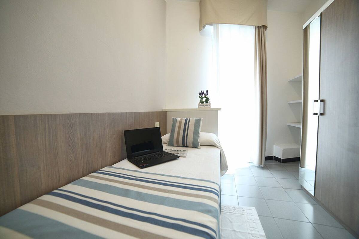 A bedroom with a bed, a laptop, a window with curtains, and a wardrobe.