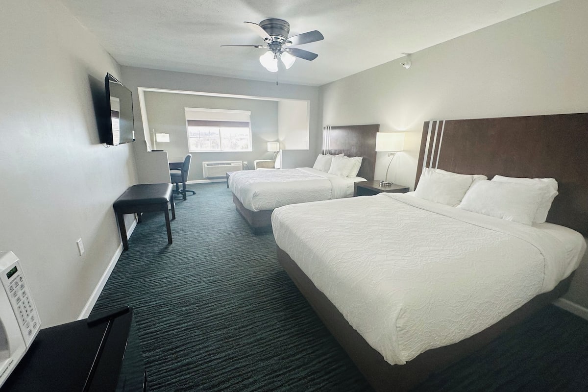 A hotel room with two beds, a desk, a chair, a ceiling fan, and a window.