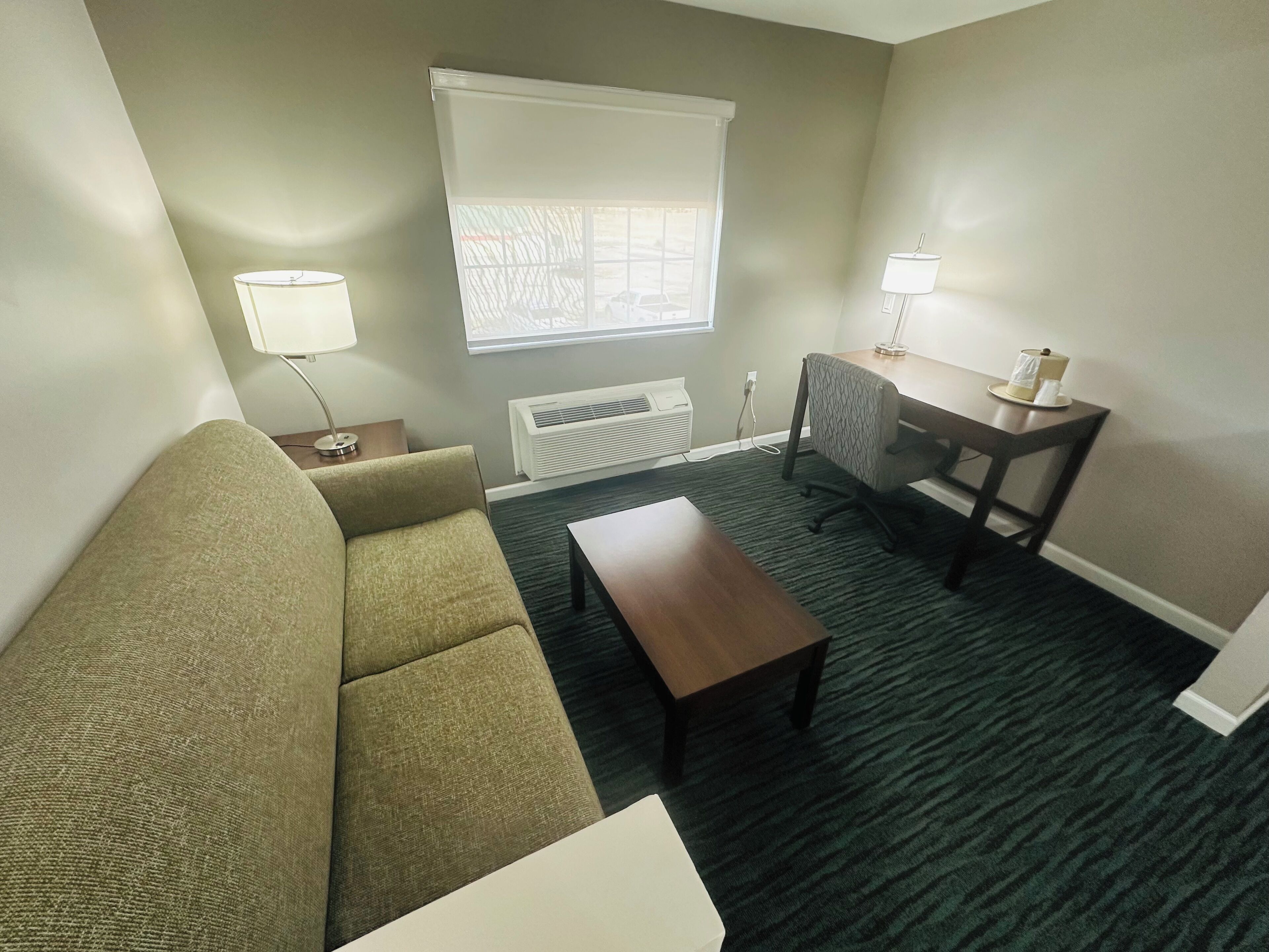 A living room with a sofa, a coffee table, a desk with a lamp, a window with blinds, and a radiator.
