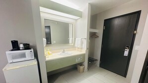 A compact hotel bathroom with a white sink, a mirror, a microwave, and a dark door.