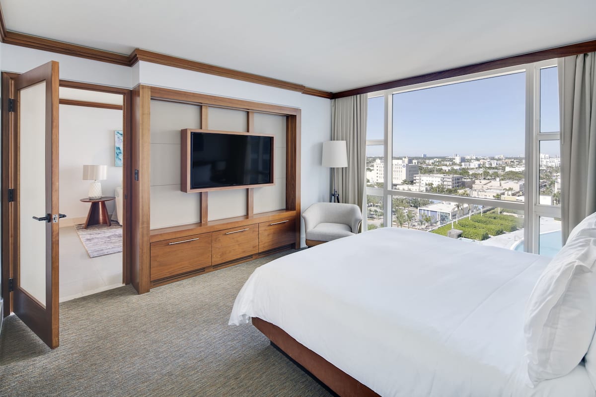 A hotel room with a large bed, a TV mounted on a wooden panel, a chair, and a view of the city through a large window.
