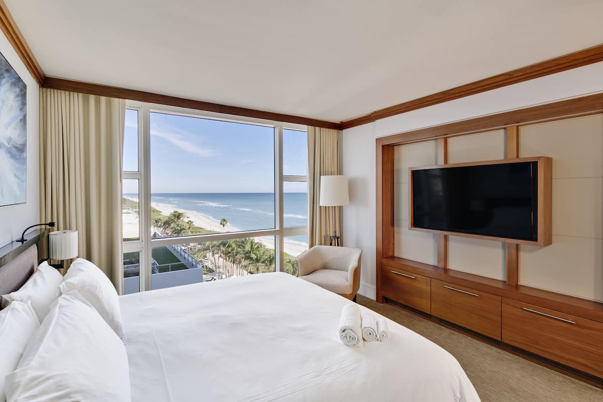 A hotel room with a large bed, a TV, a chair, and a view of the ocean.