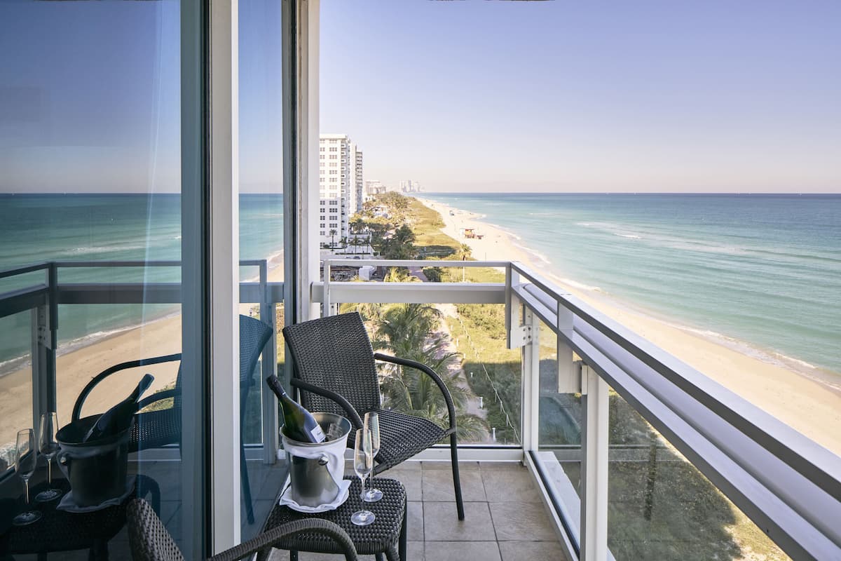 A balcony with a view of the beach and ocean, a chair, a bottle of champagne, and a glass.