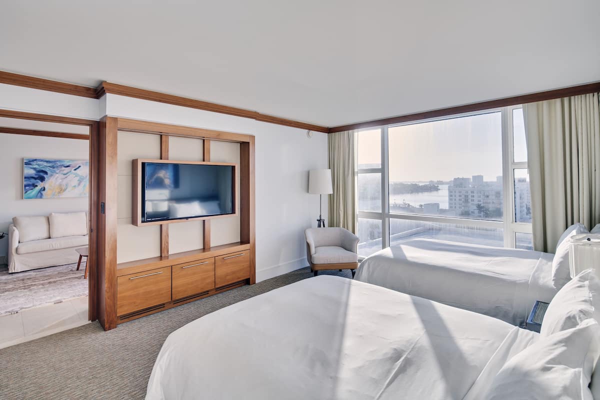 A hotel room with a large bed, a TV mounted on a wooden panel, a sofa, and a view of the city through large windows.