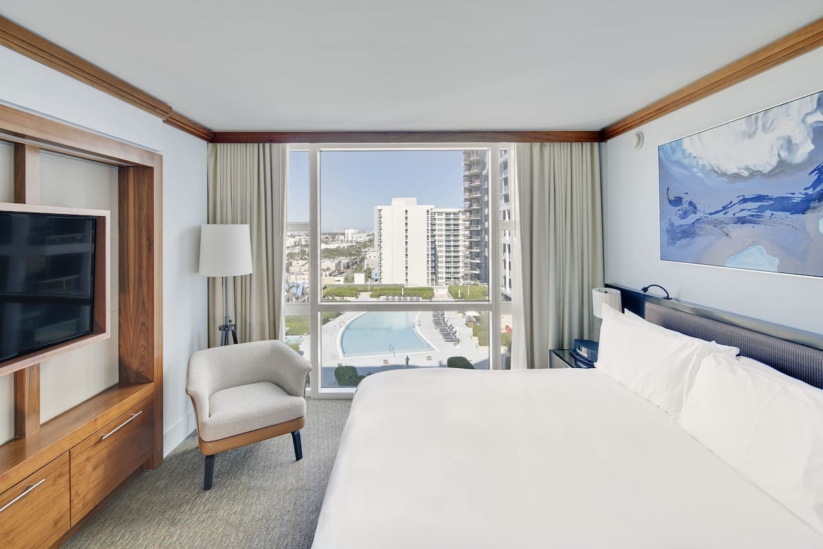 A hotel room with a large bed, a TV, a chair, and a view of a swimming pool and buildings.