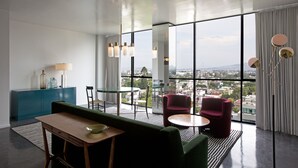 Une chambre d’hôtel moderne dotée d’une grande fenêtre donnant sur la ville, d’un canapé vert, de deux fauteuils rouges, d’une petite table ronde et d’un meuble bleu.