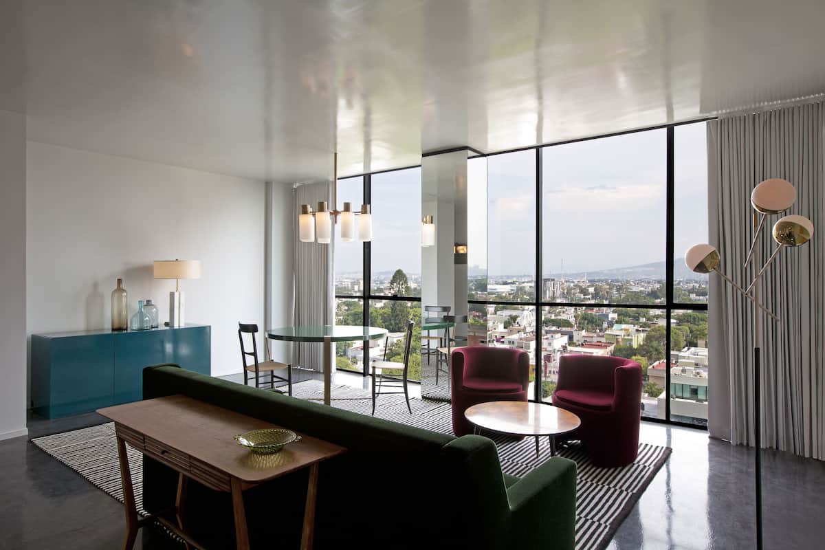 A modern hotel room with a large window offering a city view, a green sofa, two red armchairs, a small round table, and a blue cabinet.