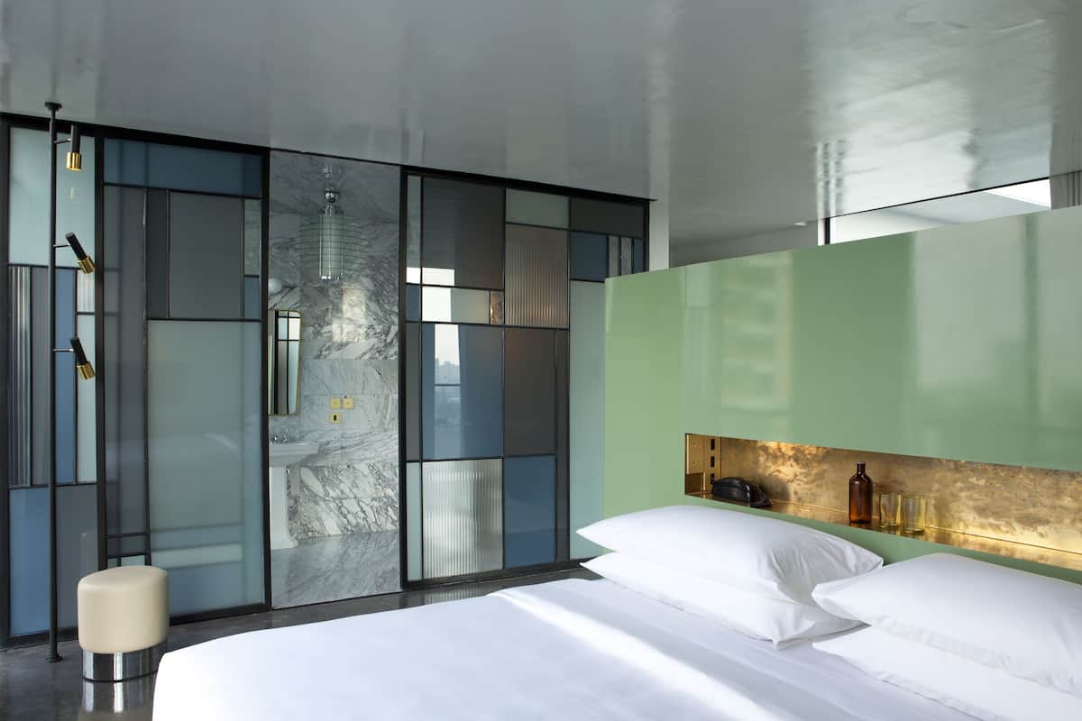 A modern hotel room with a large bed, a glass partition wall, and a green accent wall.