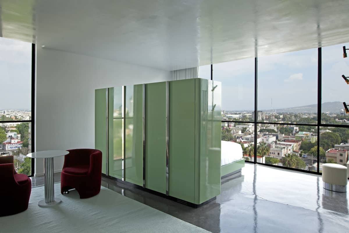 A modern hotel room with large windows, a red chair, a small table, and green cabinets.