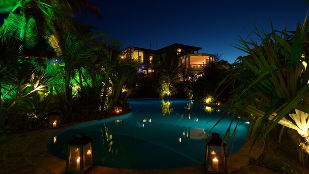 Vista nocturna de un área de piscina con una casa al fondo, rodeada de palmeras e iluminada por faroles.