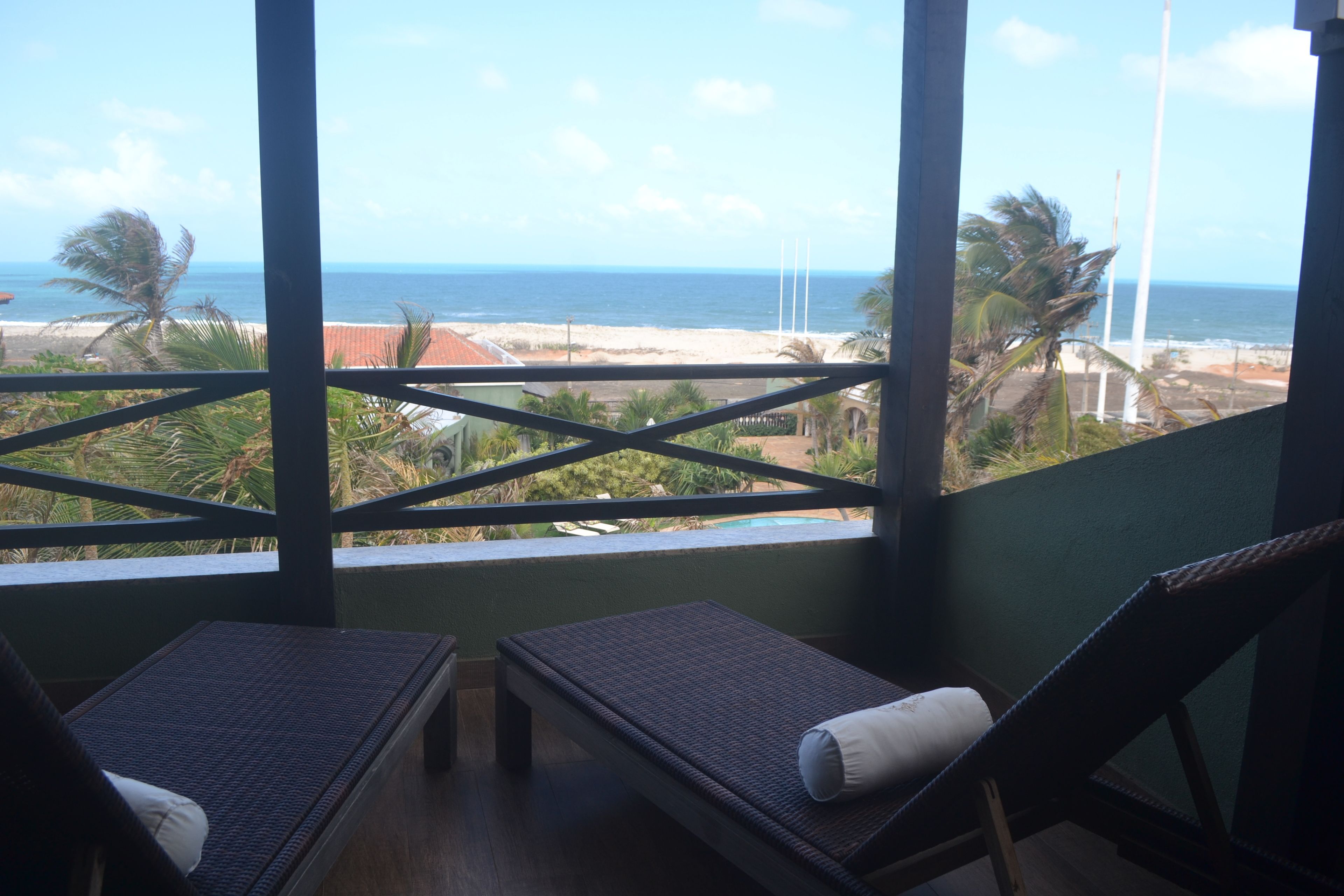 Quarto com duas poltronas de vime, vista para a praia e oceano, e uma grade com palmeiras.
