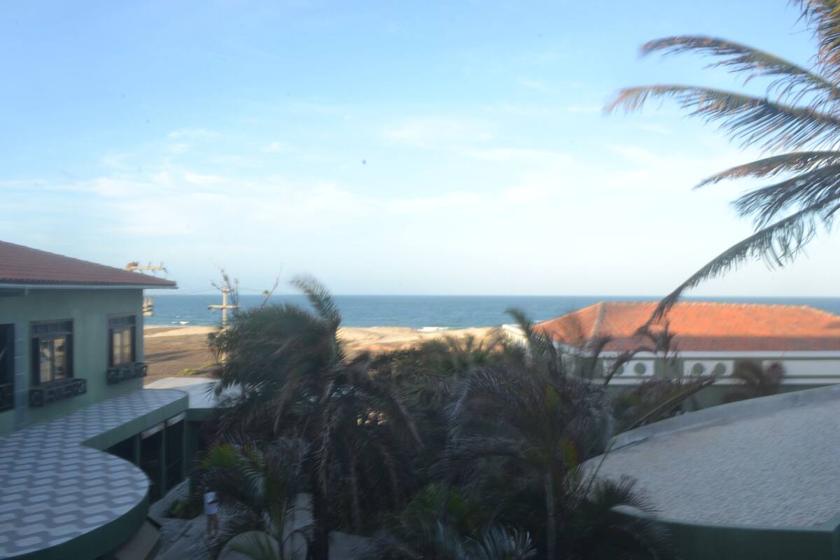 Vista de una playa con un edificio al frente, una palmera y el océano de fondo.