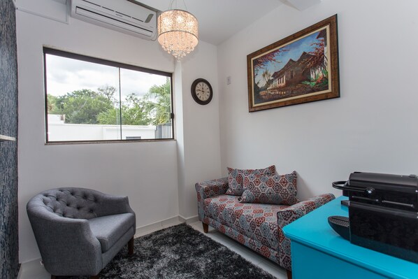 Una habitación con un sillón gris, un sofá estampado, una mesa azul, un cuadro y un reloj en la pared.
