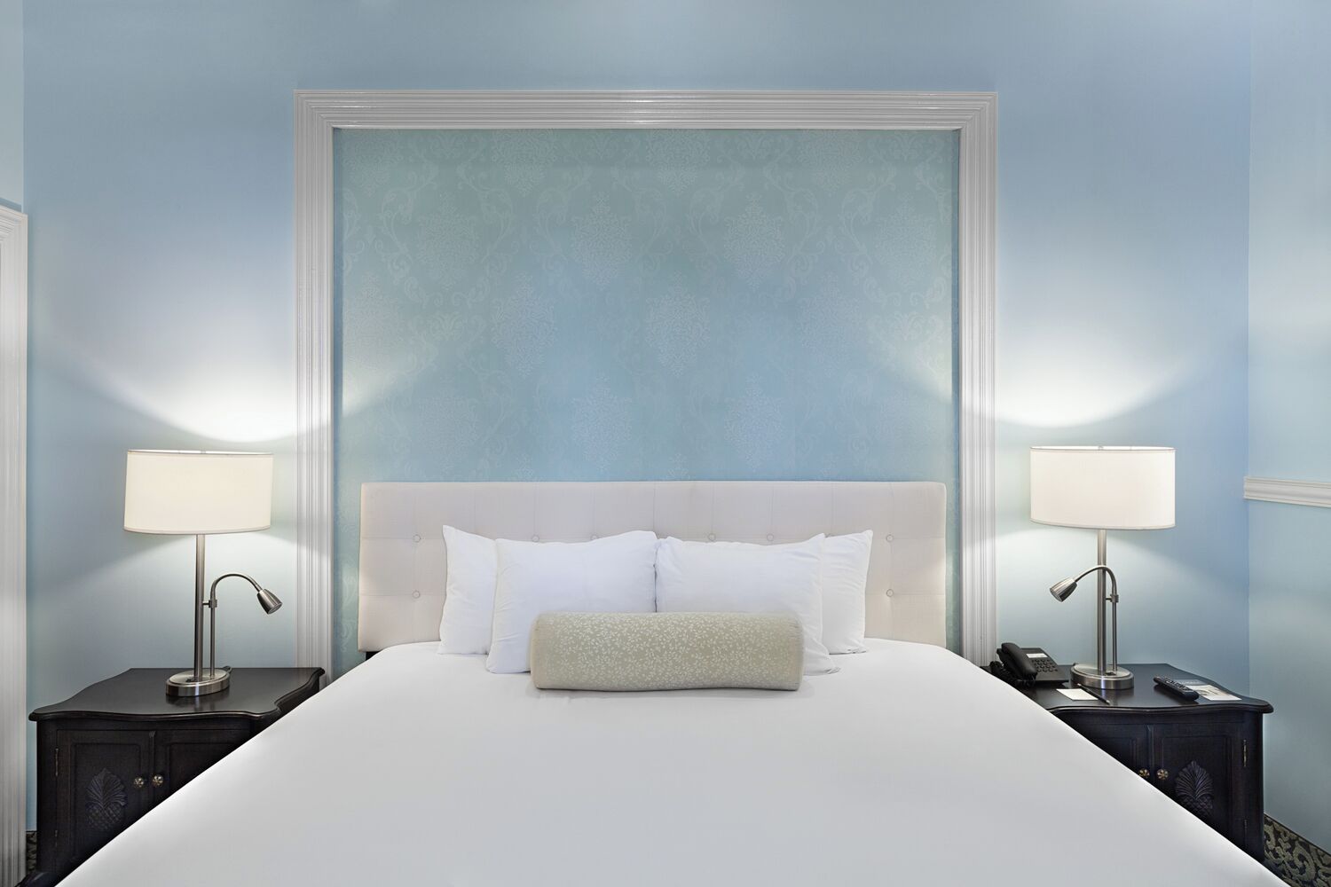 Una cama bien hecha con sábanas blancas y un cojín de color beige, flanqueada por dos mesitas de noche con lámparas, en una habitación con un cabecero grande y ornamentado y paredes azul claro.