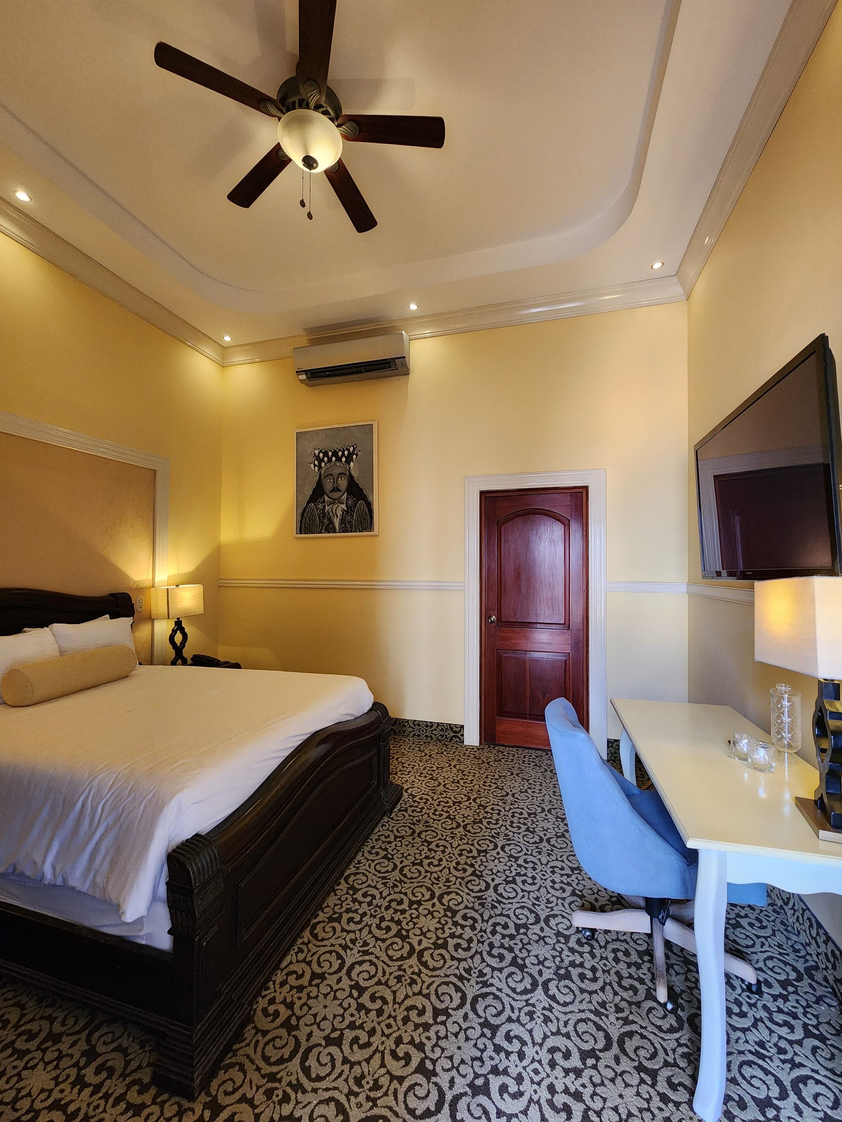 A hotel room with a bed, a desk, a chair, a ceiling fan, a TV, and a door.