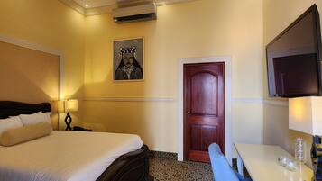 A hotel room with a bed, a desk, a chair, a ceiling fan, a TV, and a door.