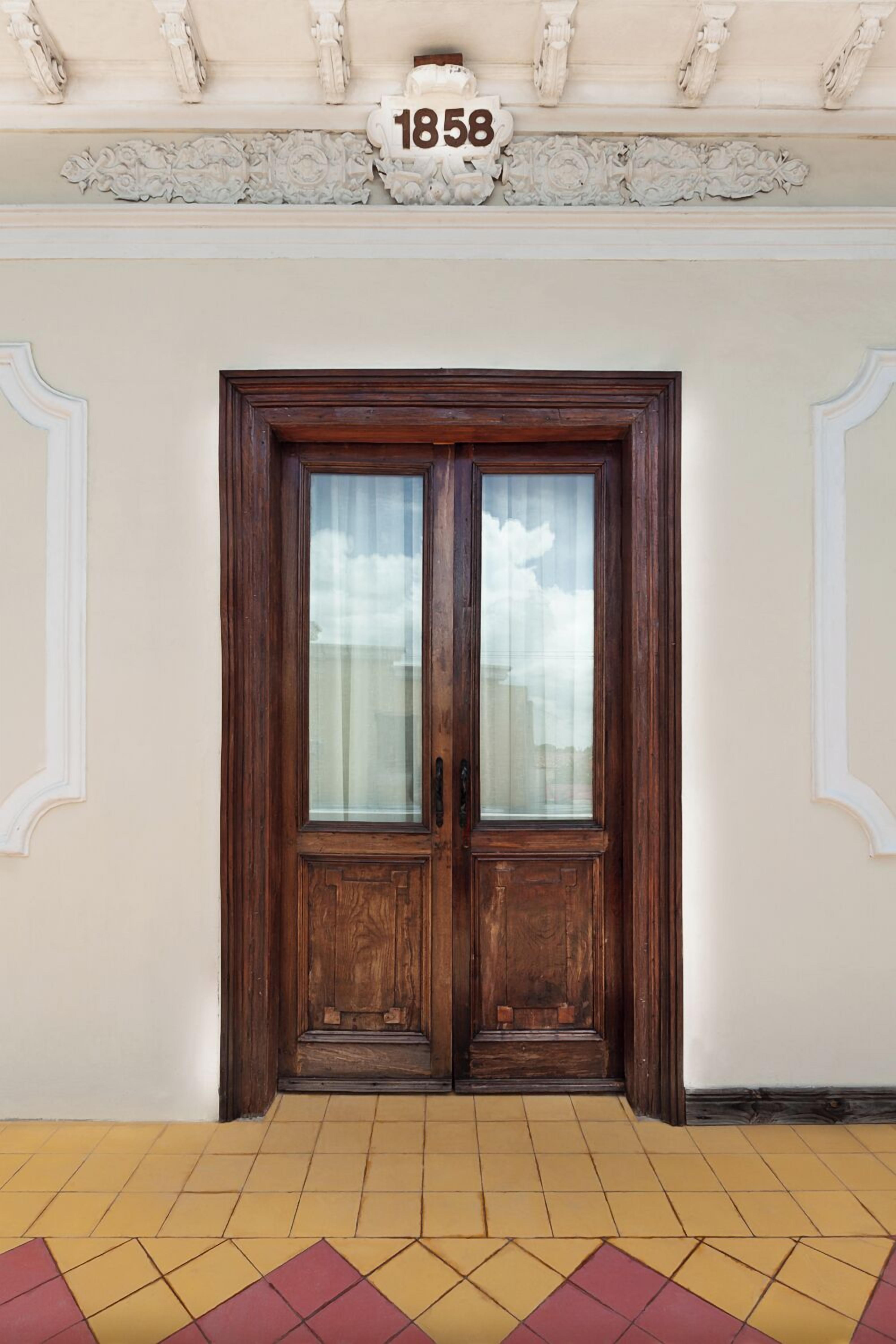 A wooden double door with a glass panel, a number plaque above it, and a yellow and red patterned floor.