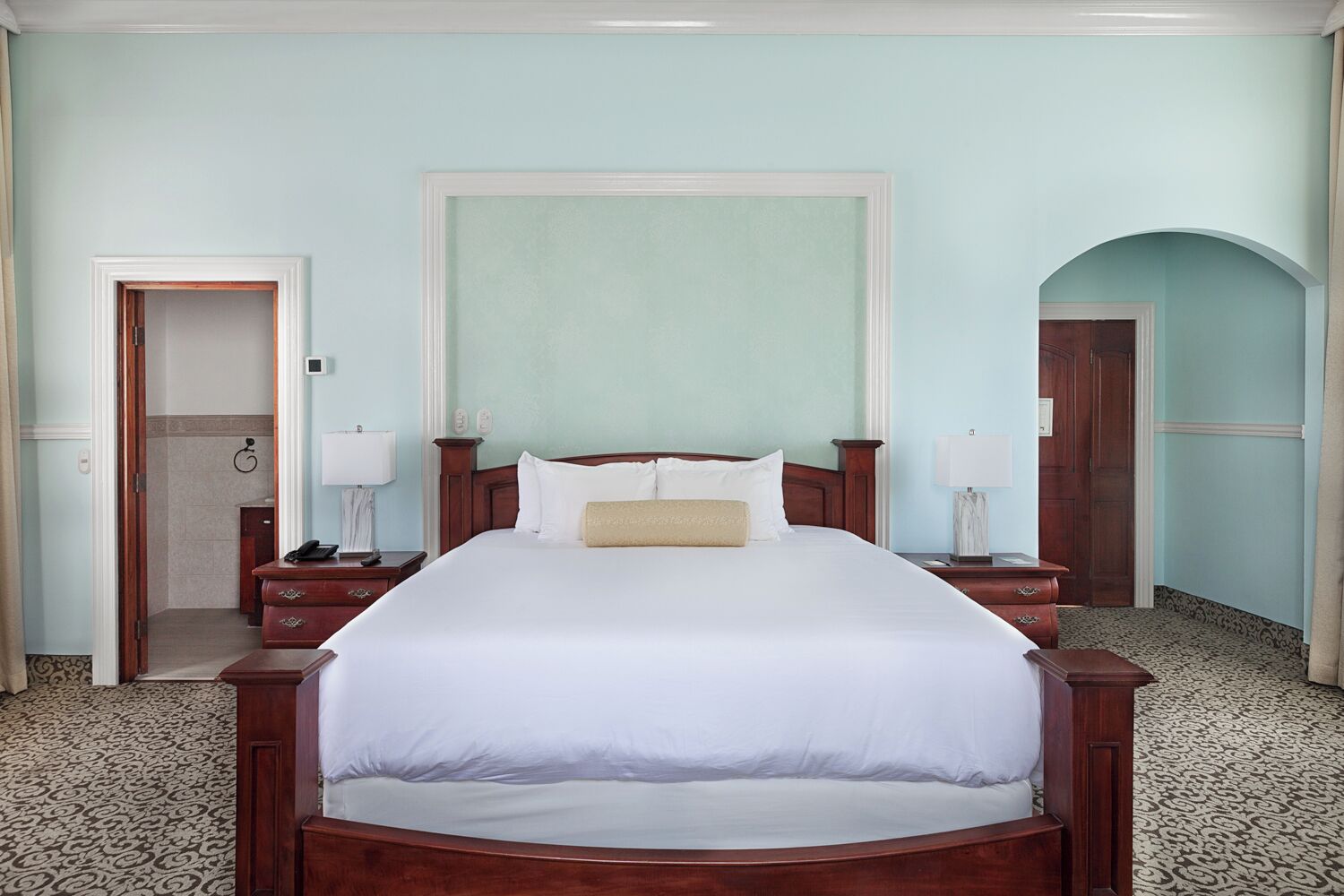 A hotel room with a large bed, two bedside tables, a nightstand, a door, and a small bathroom.
