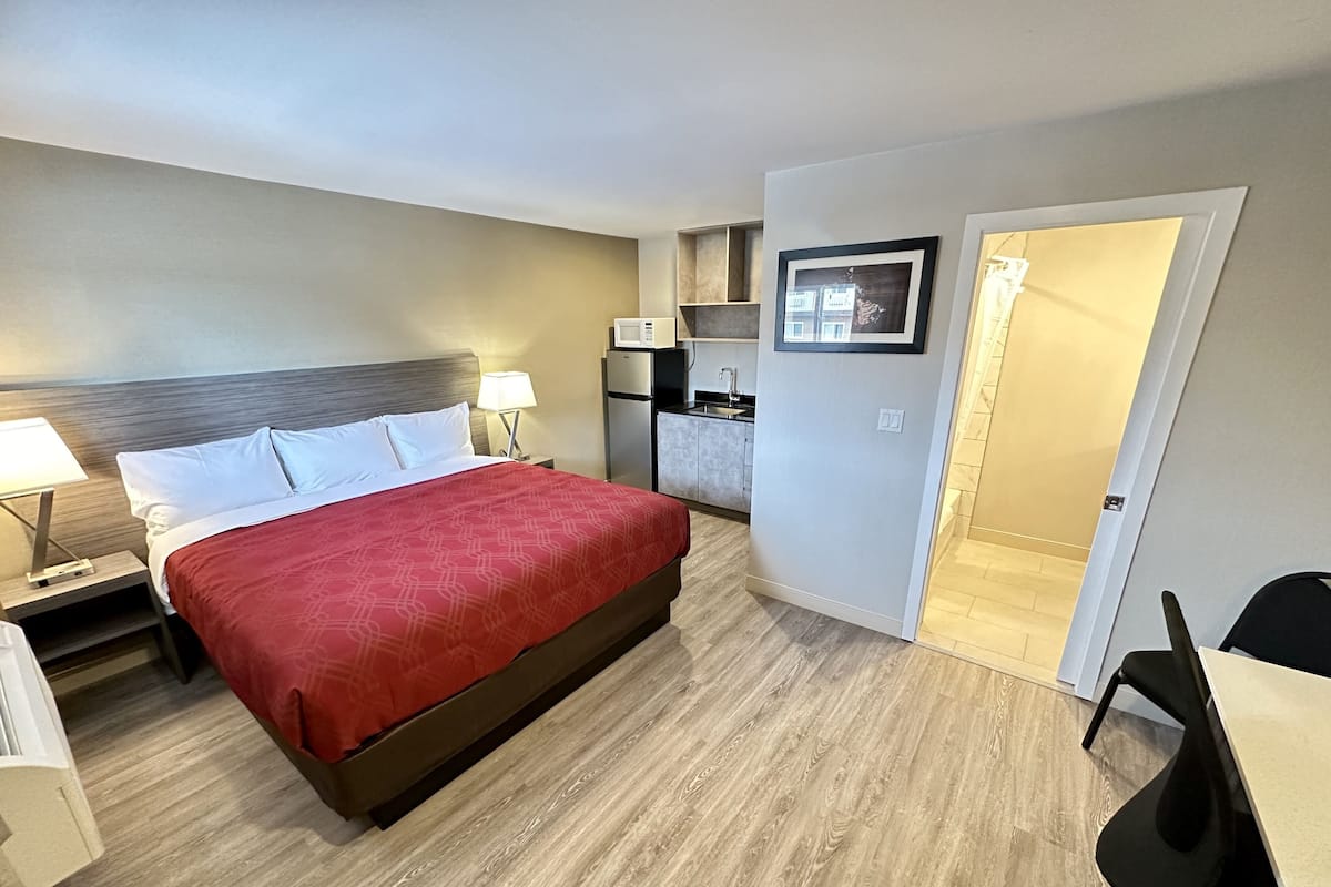 A hotel room with a bed, a nightstand, a desk, a chair, a kitchenette, and a bathroom.