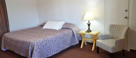 A hotel room with a bed, a chair, a nightstand with a lamp, and a door.