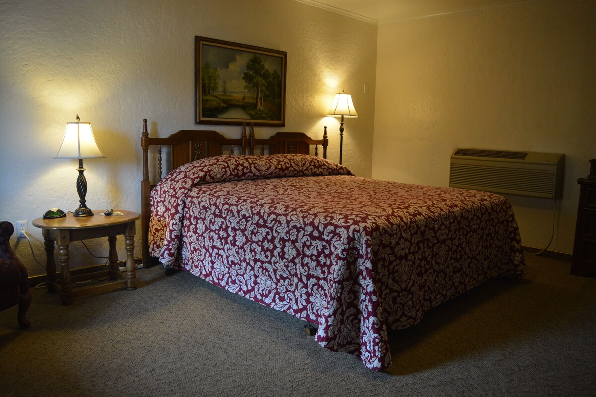 A bedroom with a bed, two bedside lamps, a painting on the wall, and an air conditioning unit.