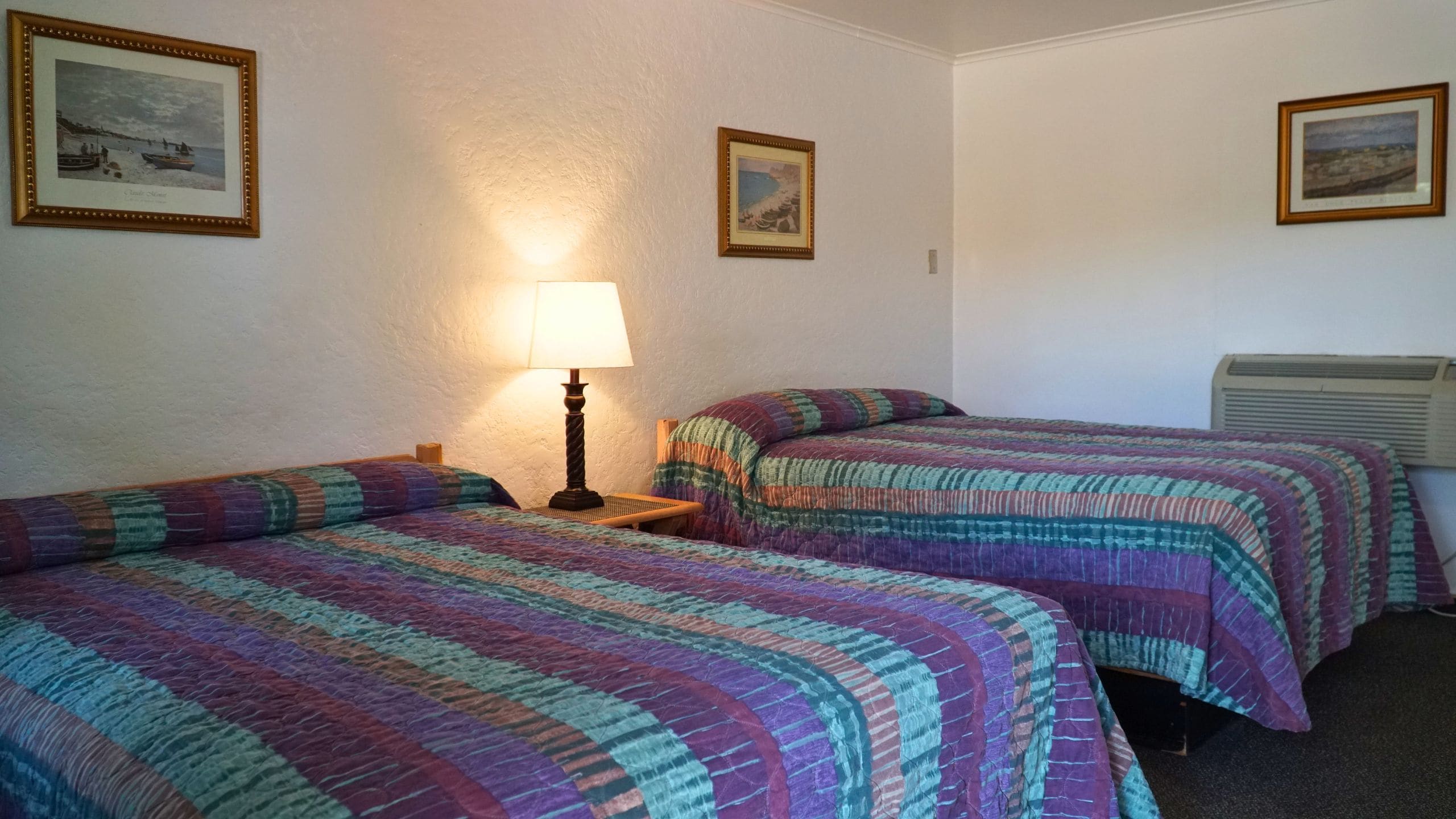 Hotellihuone, jossa on kaksi sänkyä, yöpöytä, lamppu ja seinillä olevia tauluja.