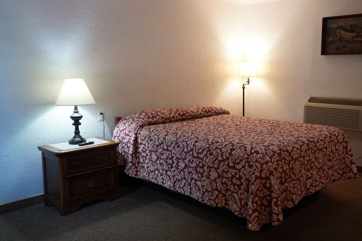 A bedroom with a bed, a nightstand, a lamp, an air conditioner, and a painting on the wall.
