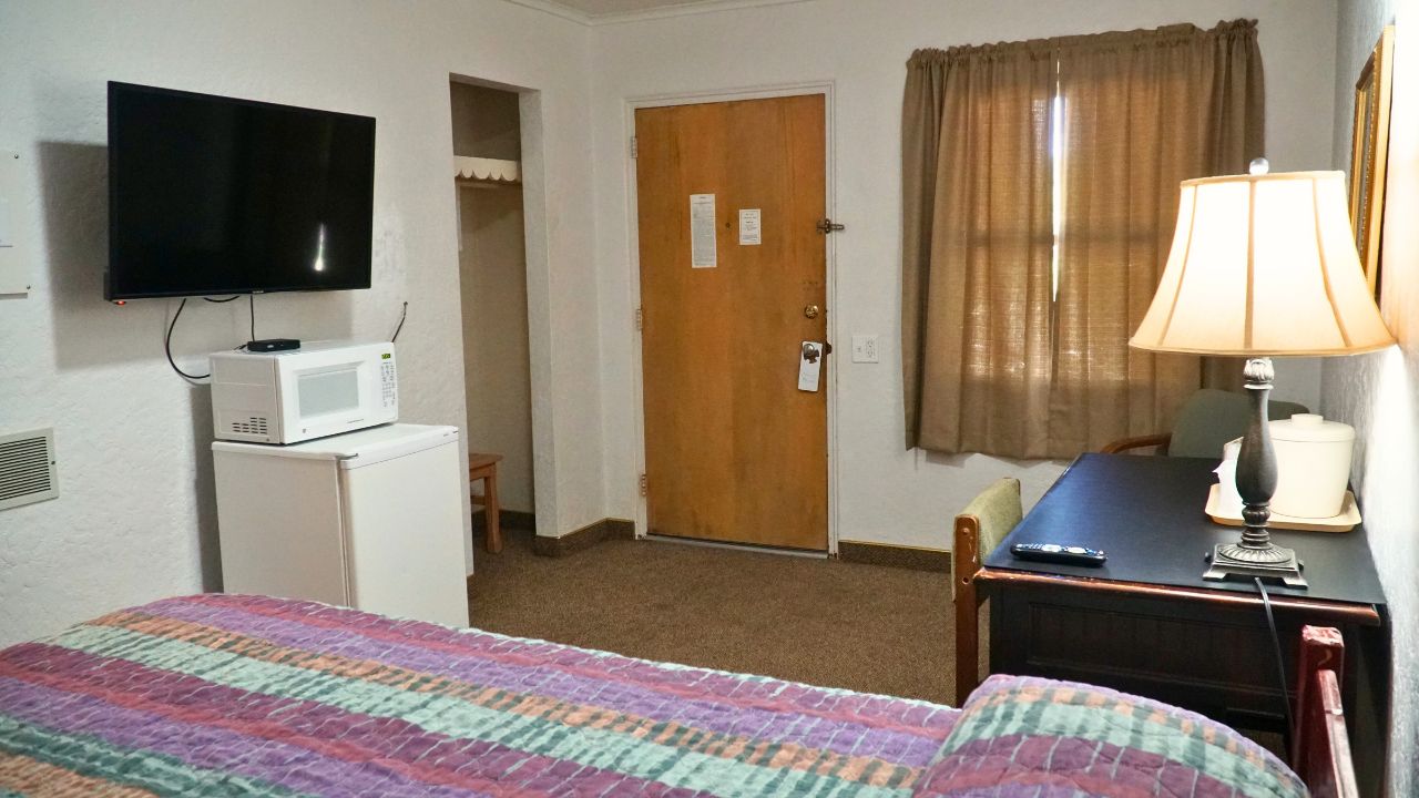 A hotel room with a bed, a desk, a television, a microwave, a door, and a window with curtains.