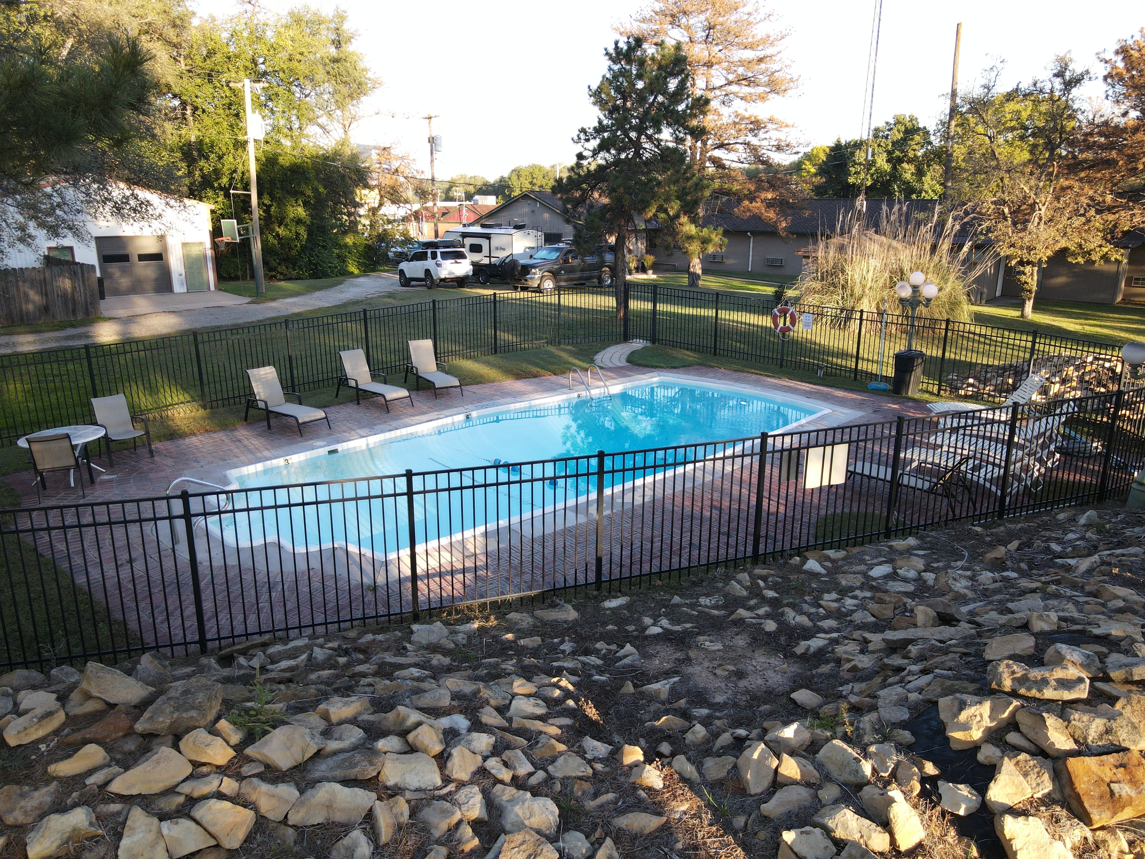 A rectangular swimming pool with a black metal fence, surrounded by a paved deck and a grassy area.