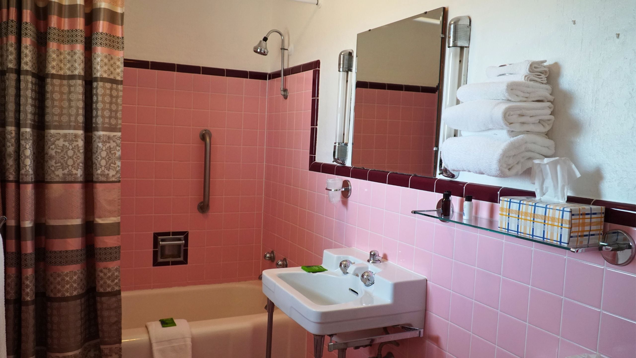 A bathroom with a pink tiled wall, a white sink, a handheld showerhead, a towel rack with folded towels, and a glass shelf with toiletries.