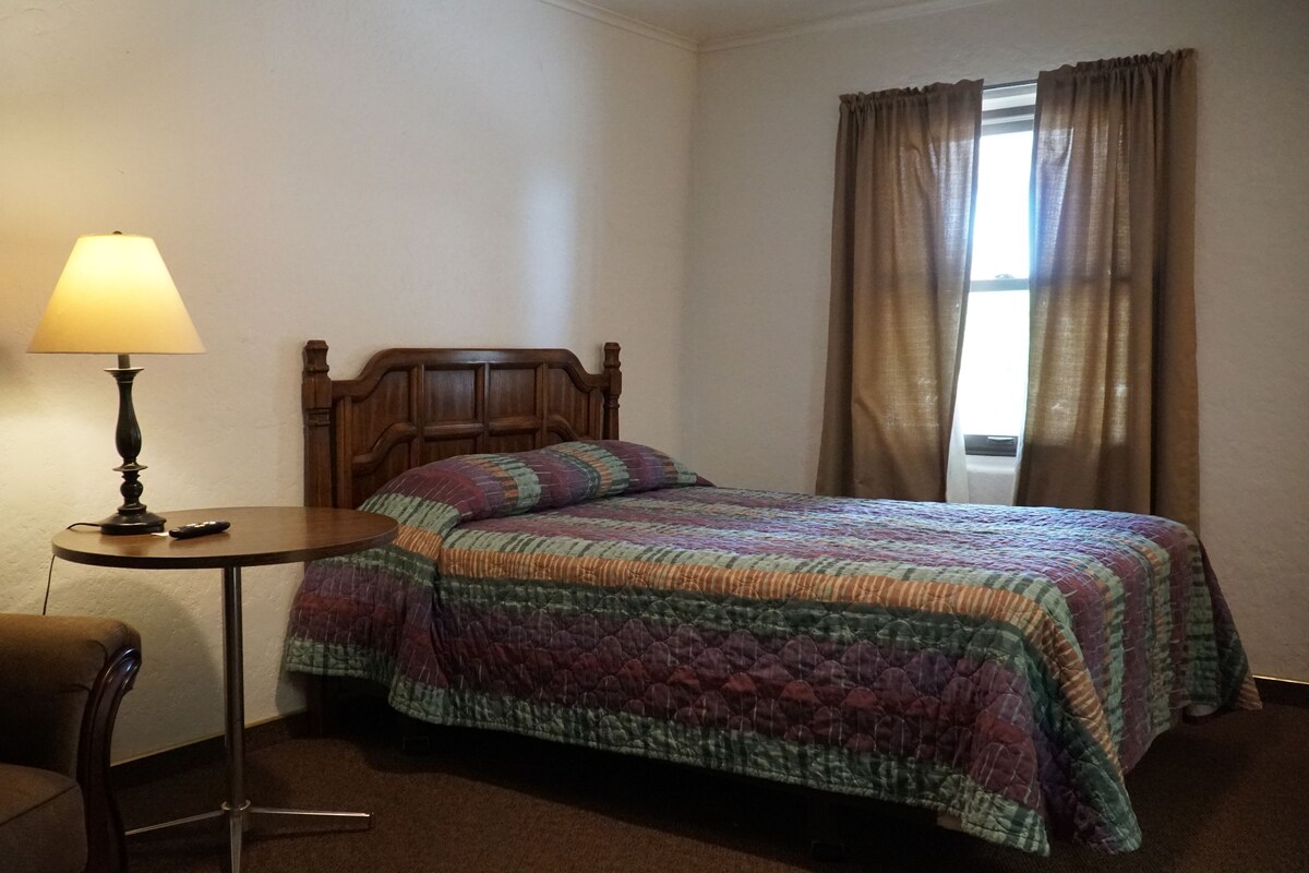 A hotel room with a bed, a bedside table with a lamp, a chair, and a window with curtains.