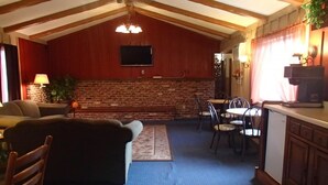 Una habitación acogedora con chimenea de ladrillo, vigas de madera en el techo, un televisor y un comedor con sillas y mesa.