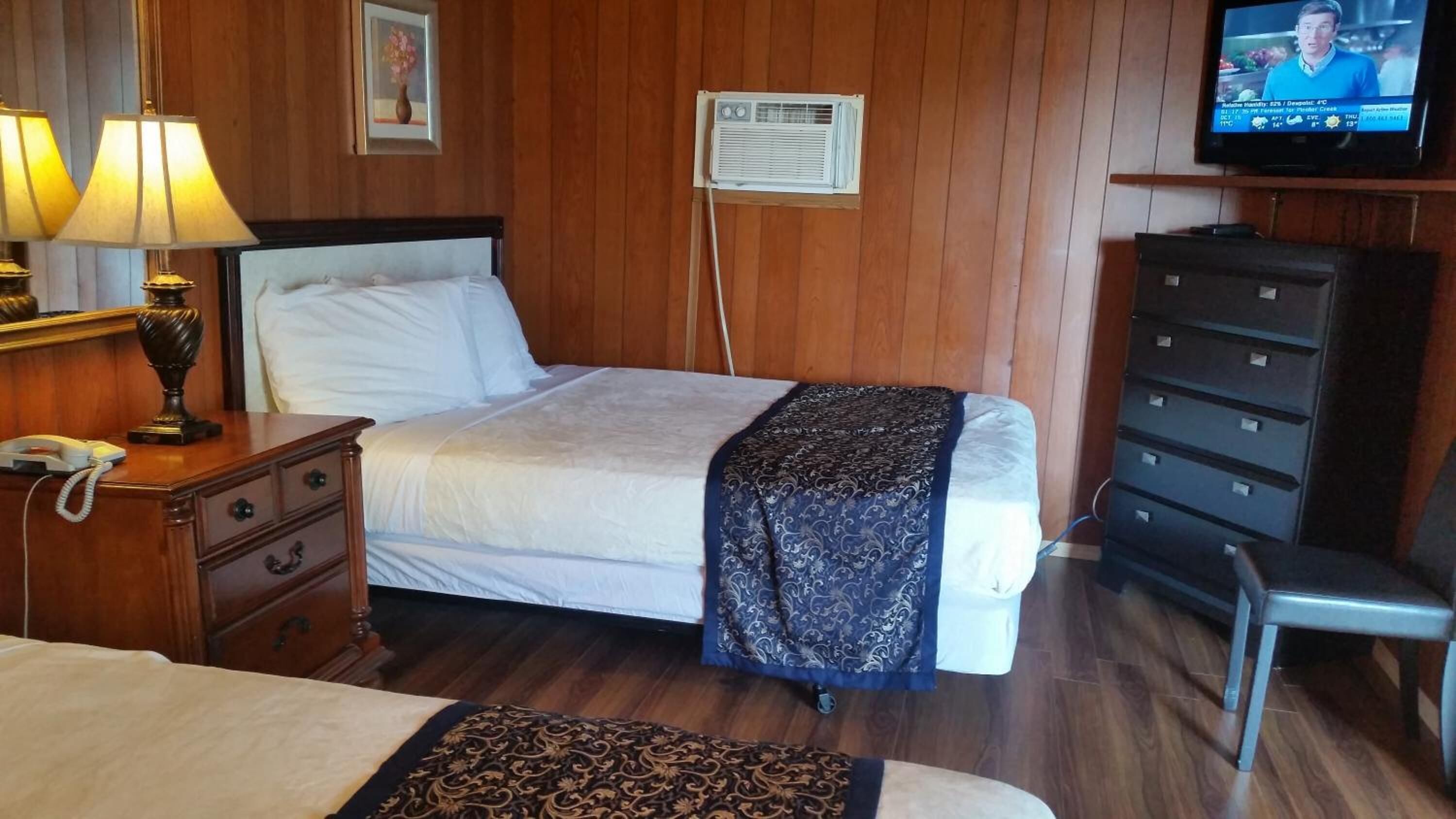 Habitación de hotel con cama, mesita de noche con lámpara, cómoda, televisor y aire acondicionado.