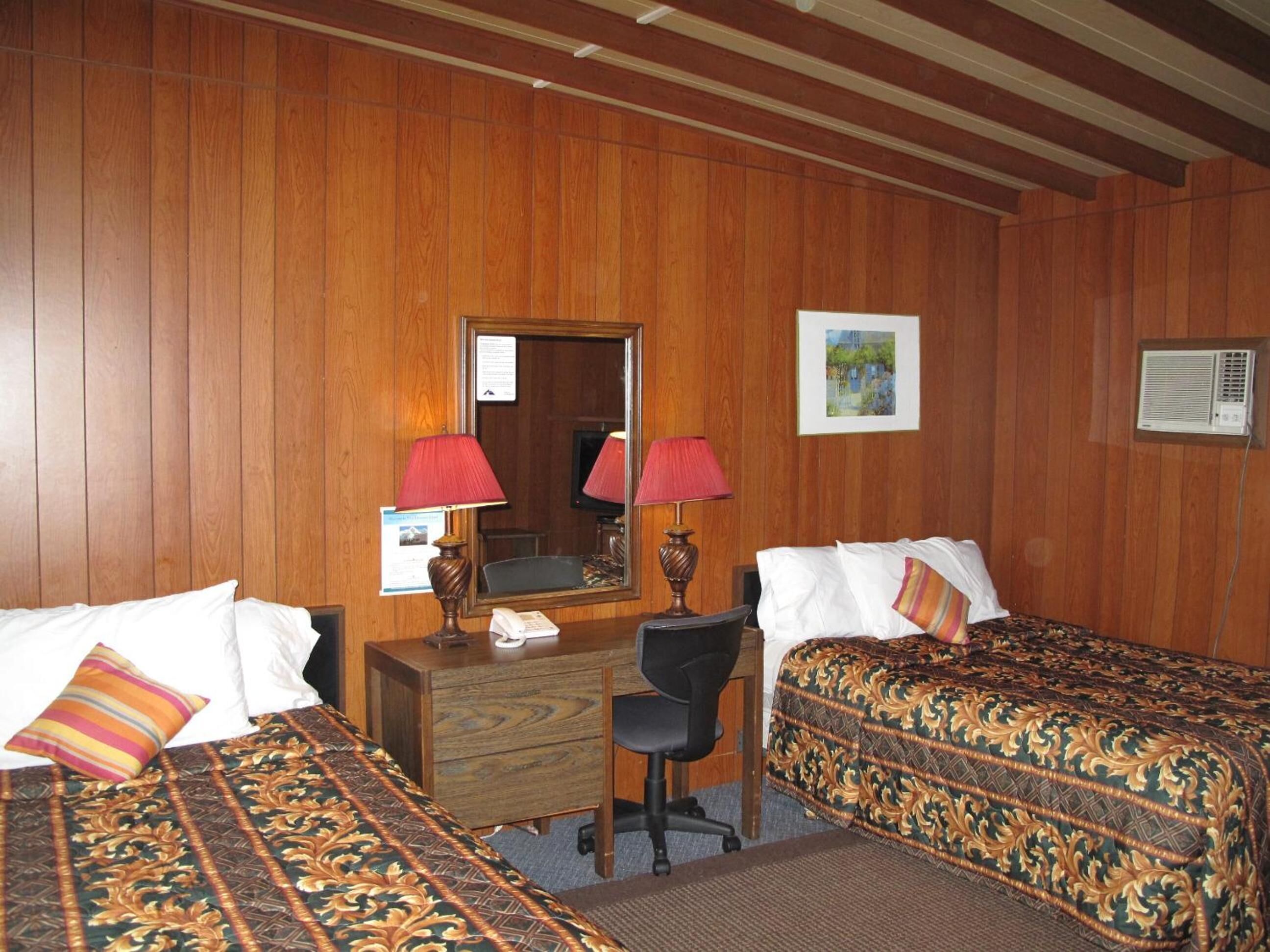 A hotel room with wooden panel walls, a desk with a mirror, two beds with patterned bedding, a chair, and a small desk lamp.