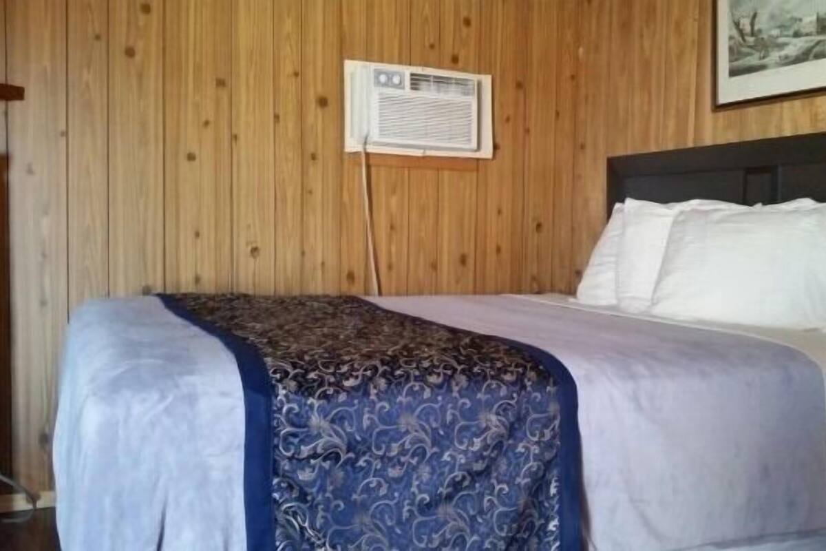 Un lit bien fait, recouvert d’une couverture à motifs et agrémenté d’oreillers blancs, un mur en bois orné d’un tableau encadré et un petit appareil de climatisation.