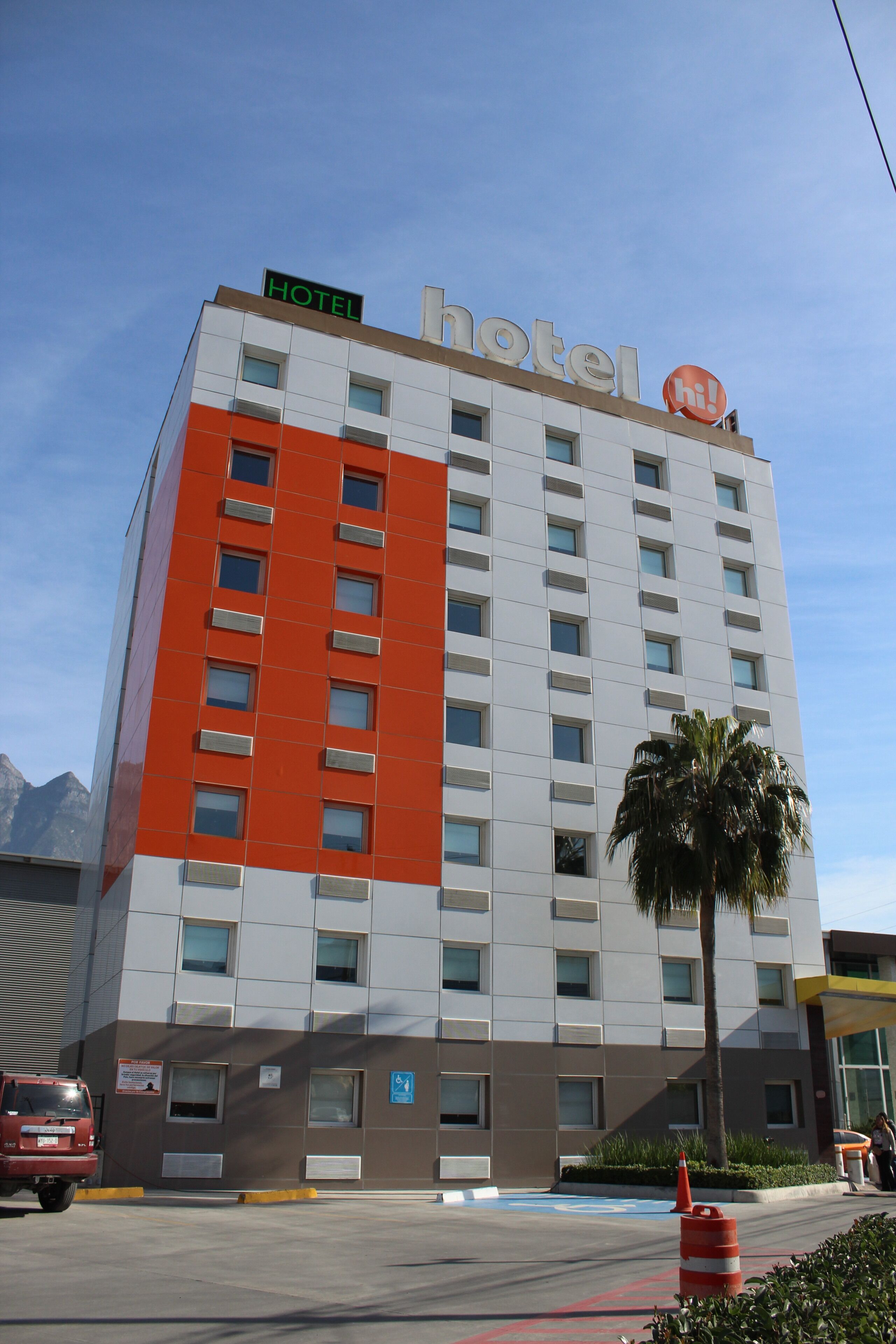 Un hôtel à plusieurs étages, avec une grande enseigne « HÔTEL » bien visible, un panneau bleu indiquant l’accessibilité aux personnes handicapées et un palmier au premier plan.