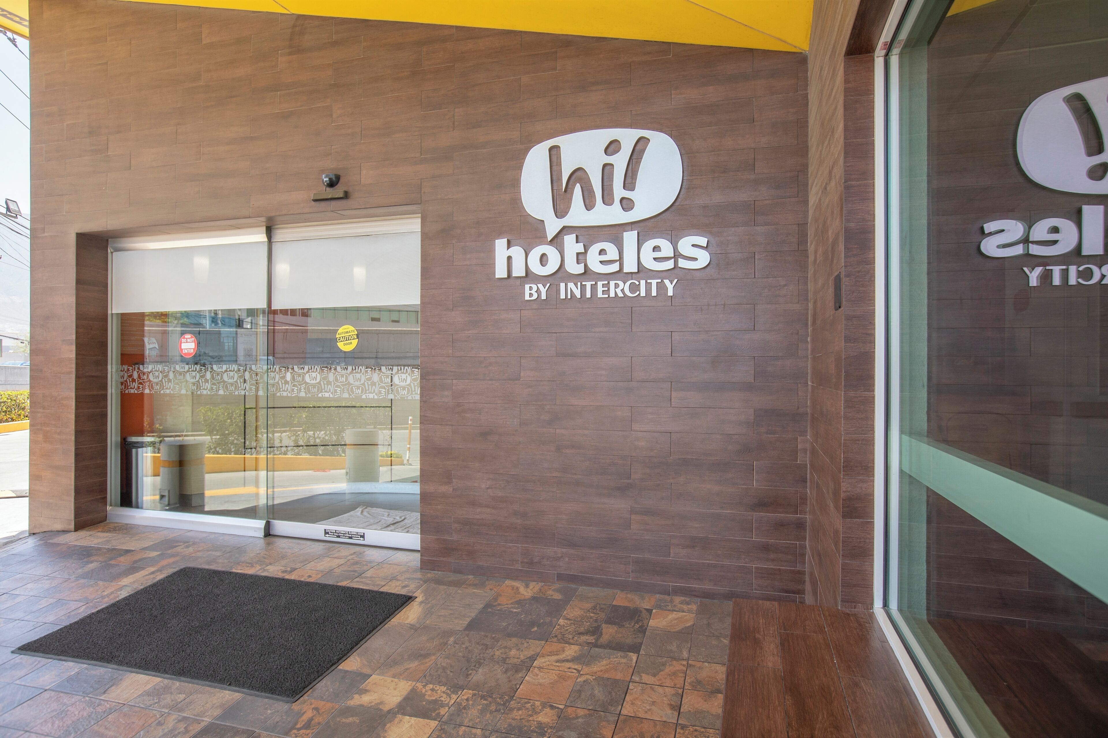 A hotel entrance with a 'hi! hoteles by Intercity' sign, a glass door, and a doormat.