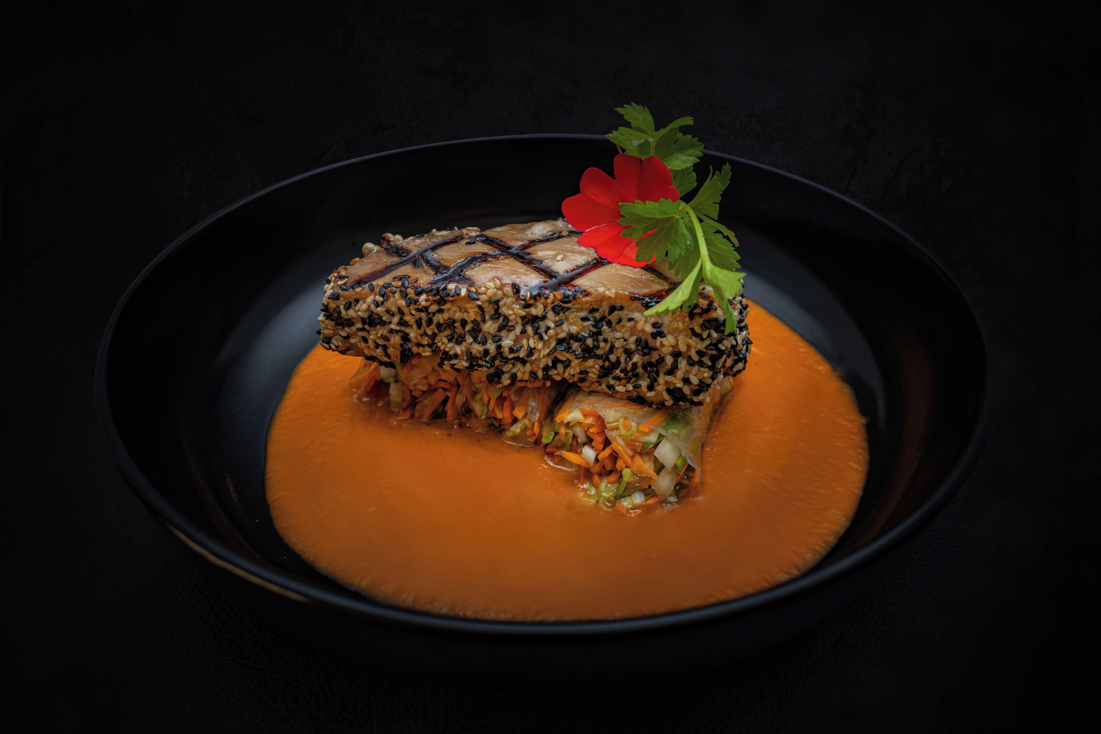A dish with a fish fillet topped with sesame seeds and a red flower garnish, served on a black plate with a smooth, orange sauce.